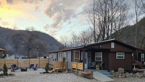 Una cabana amb un camí de grava, zona d'estar a l'exterior, una zona de foc i un paisatge de muntanya de fons.