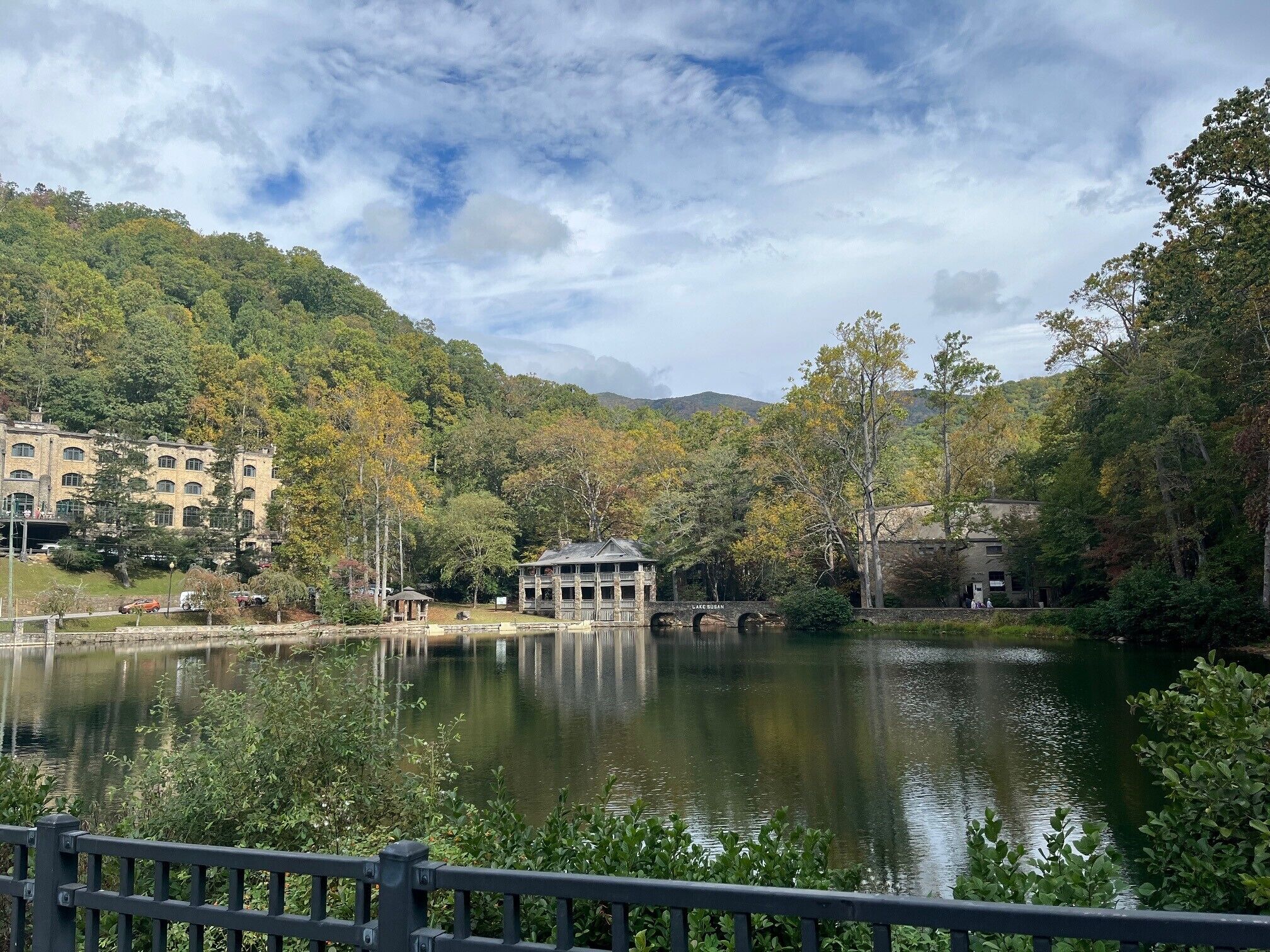 A serene lakeside view with a bridge, a building with multiple windows, and lush greenery.