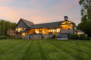 A large, modern house with a stone foundation, a prominent chimney, and a well-lit interior.