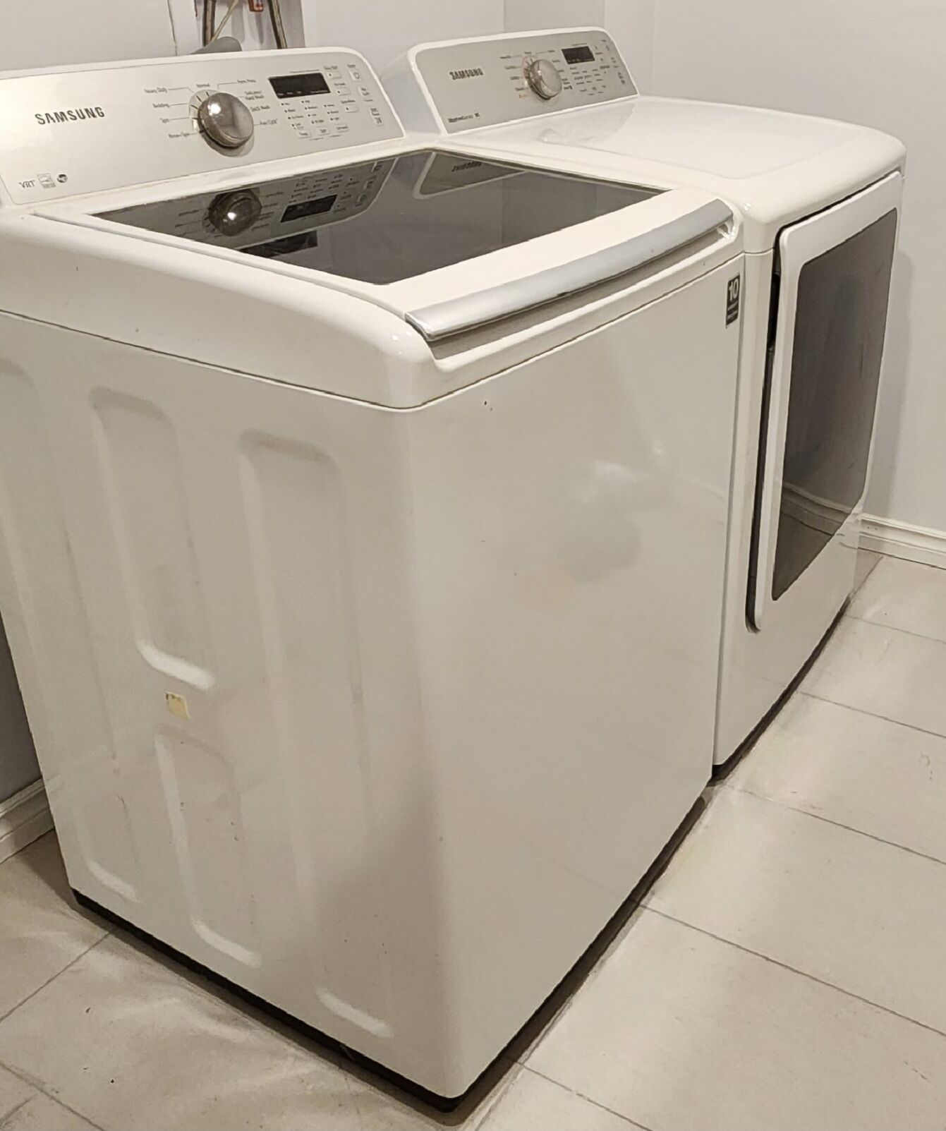 Máquina de lavar e secar Samsung, ambas com abertura frontal, numa lavanderia com chão em azulejo.