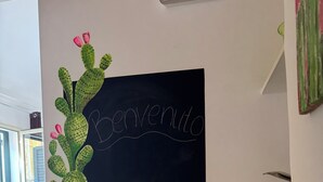 Ein Esstisch ist mit Tellern, Besteck, einer Weinflasche und Gebäck gedeckt. An der Wand befinden sich eine Kakteendekoration und eine Kreidetafel mit 'Bienvenue'.