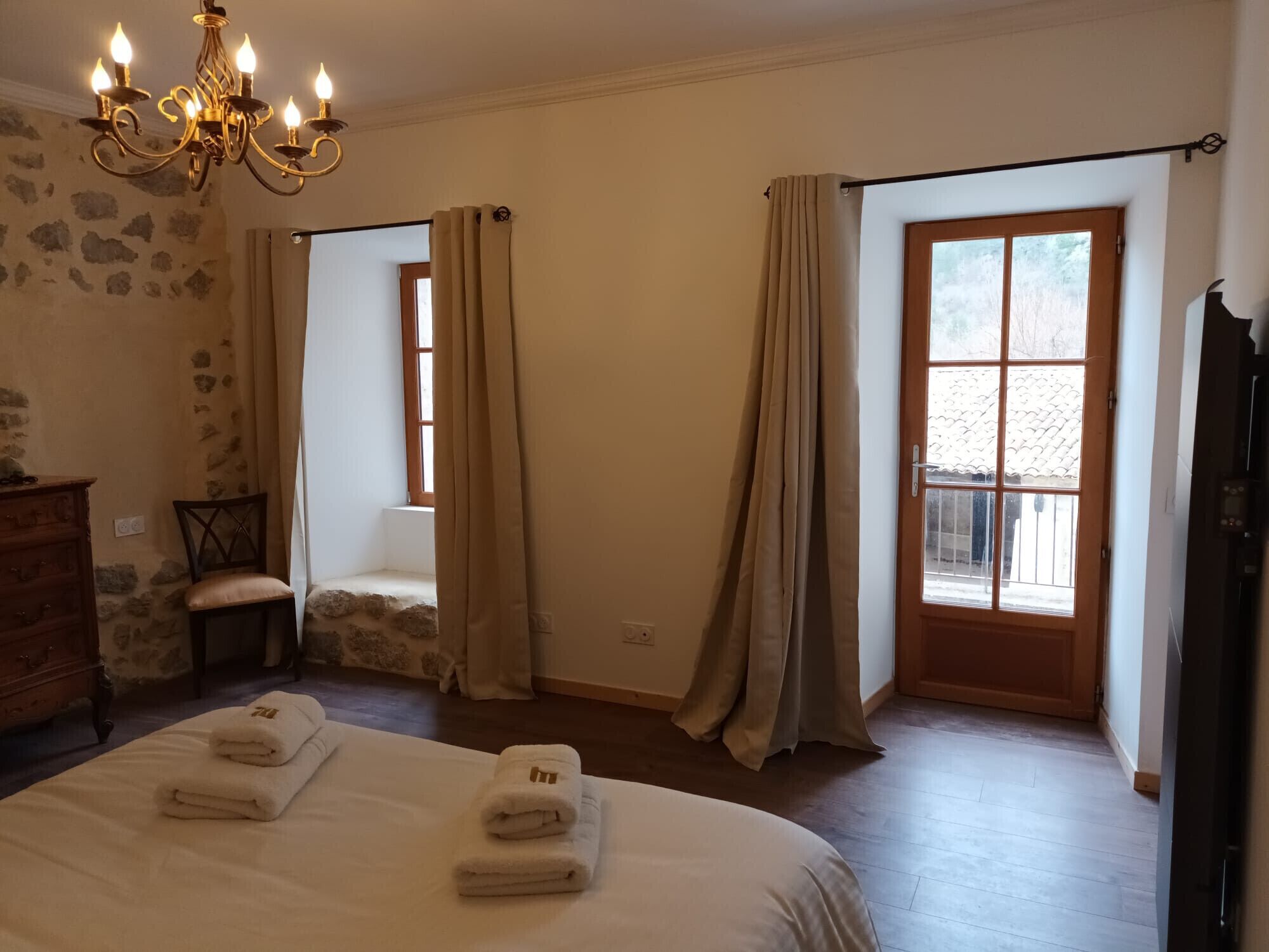 A bedroom with a bed, a wooden dresser, a chair, a chandelier, and a window with curtains.