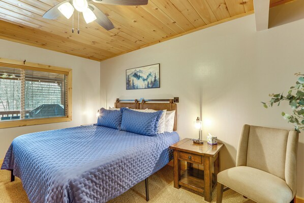 A bedroom with a bed, a nightstand, a chair, a window with blinds, a ceiling fan, and a painting on the wall. - < 1 Mi to Lake Blue Ridge: Forest-View Gem w/ Deck (Morganton)