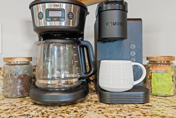 Een koffiezetapparaat, een Keurig koffiezetapparaat, een glazen kan, een witte mok en twee kruidenpotten op een aanrecht.