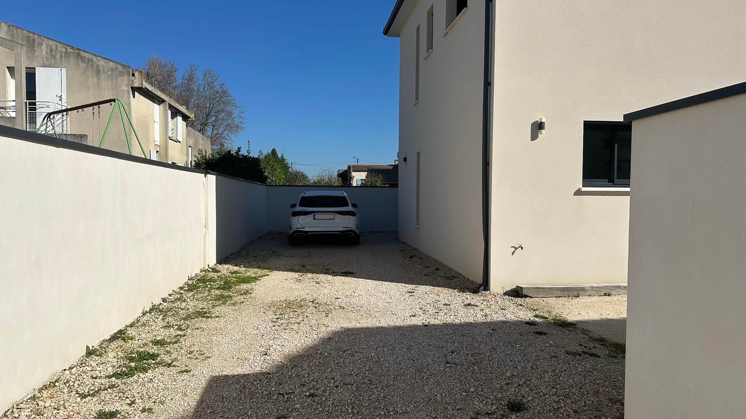Kapea soratie johtaa valkoiseen autoon, joka on pysäköity asuinalueelle. Toisella puolella tietä on rakennus ja toisella aita.