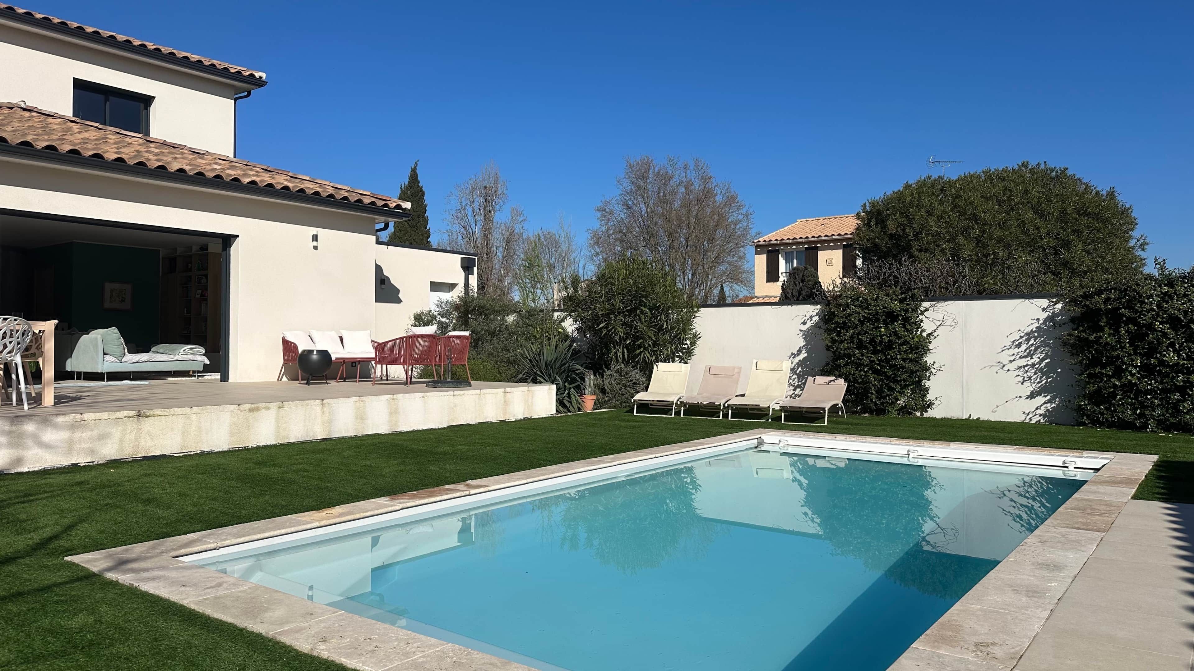 Una piscina con solárium empedrado, una zona de estar al aire libre techada y un césped bien cuidado.