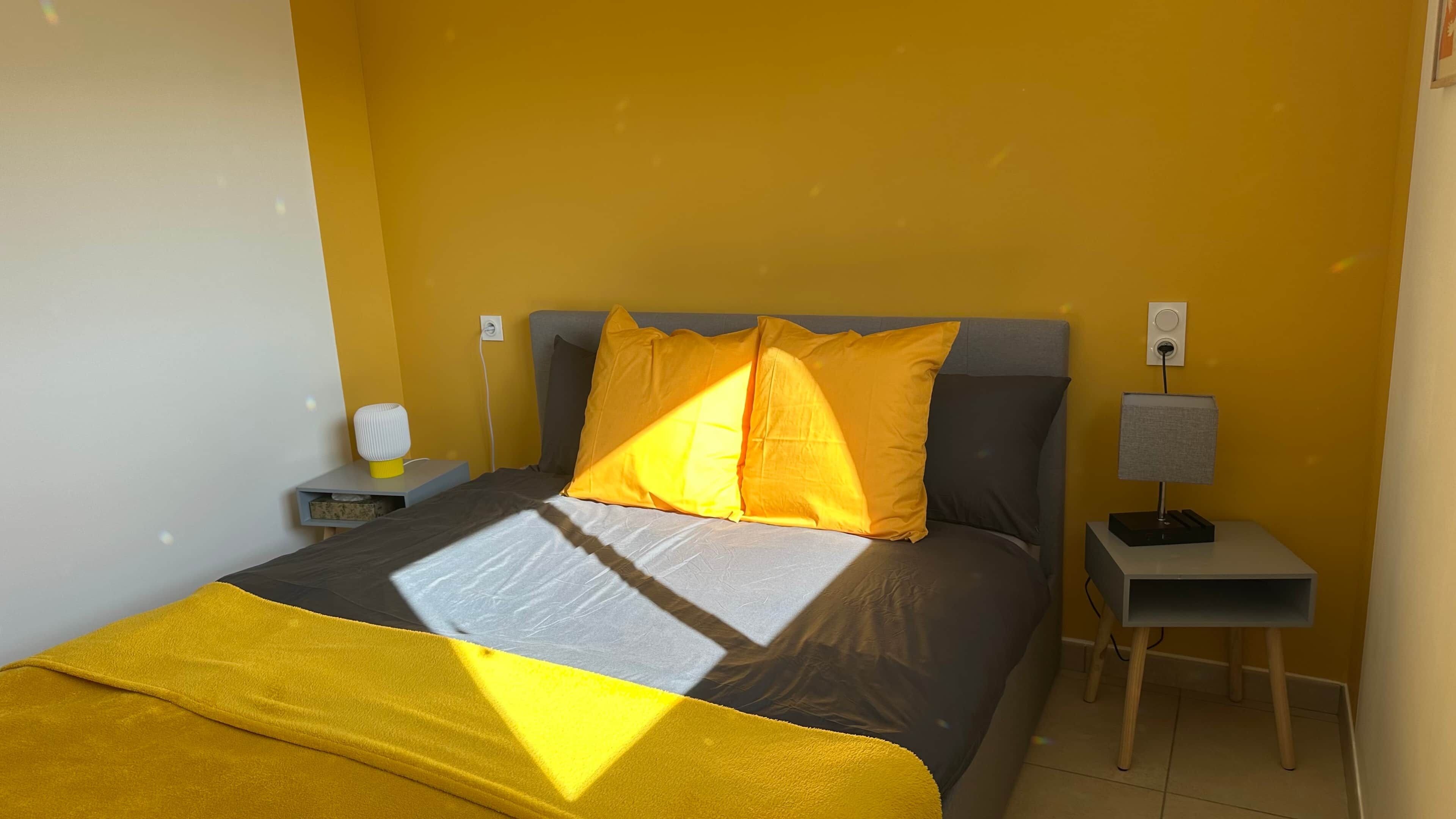 Ein Bett mit grauem Kopfteil, zwei gelben Kissen und einer graugelben Bettdecke. Ein Nachttisch mit einer Lampe und einem kleinen, weißen Gerät steht daneben. Die Wände sind gelb.