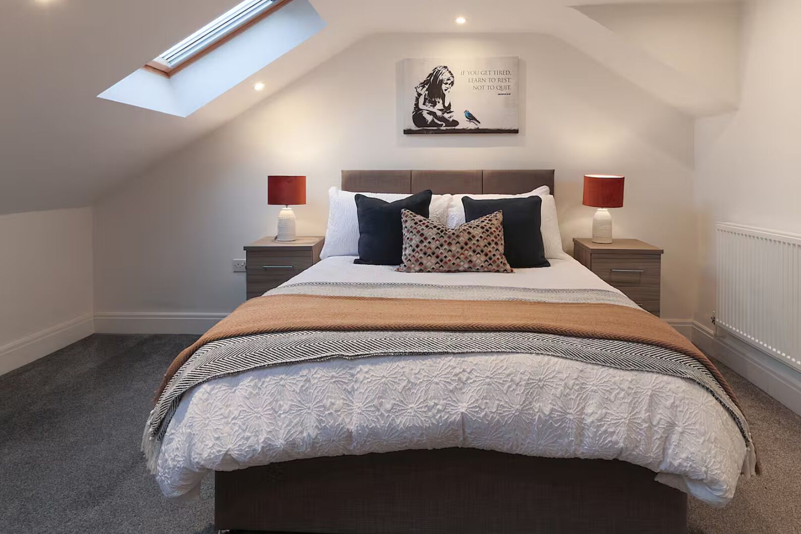 A bedroom with a skylight, a bed with multiple pillows, bedside lamps, and a motivational quote on the wall.