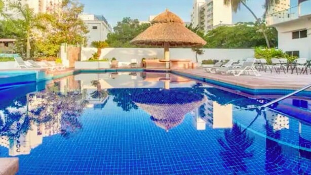 Une piscine dotée d’une structure de toit en chaume, entourée de chaises longues et d’un jardin.