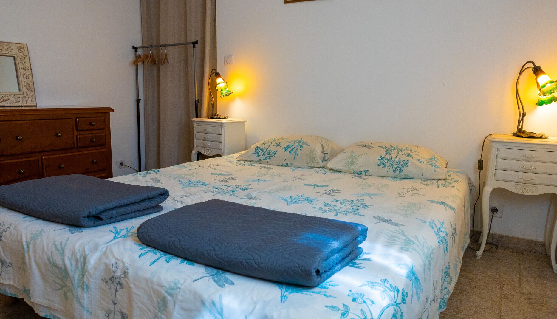 Een netjes opgemaakt bed met een bloemenbedekking, twee nachtlampjes, een houten dressoir en een nachtkastje met een lamp.