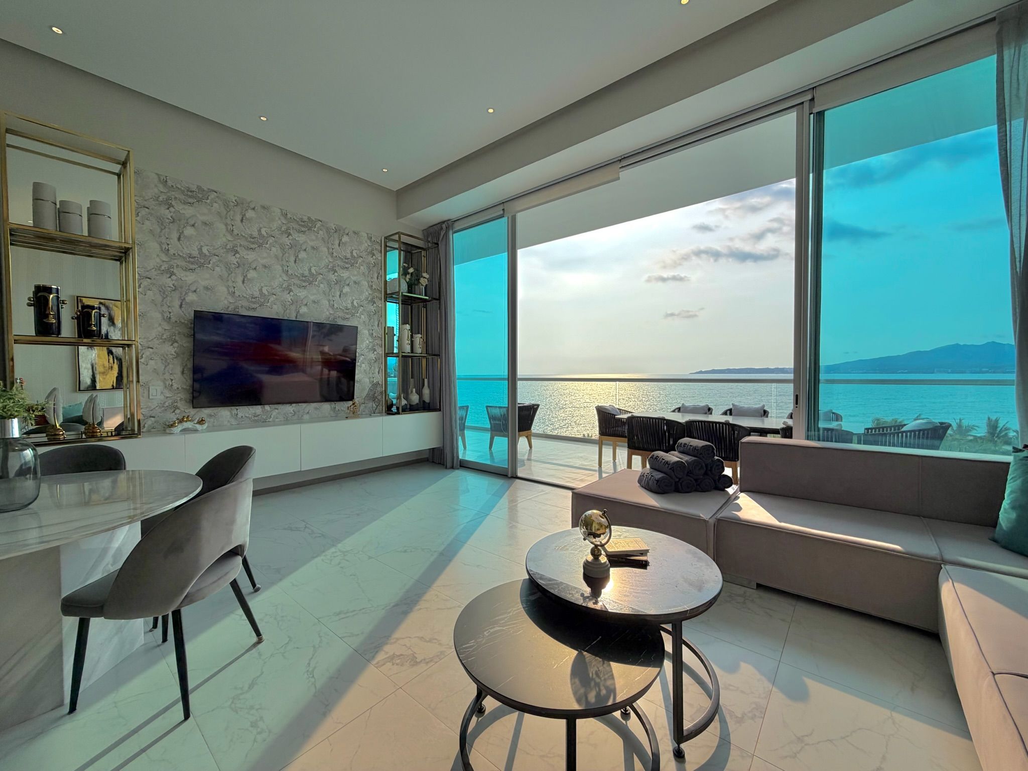Ein modernes Wohnzimmer mit großem Fenster mit Blick auf ein Gewässer, einem Essbereich mit rundem Tisch und einer Sitzgruppe bestehend aus einer Couch und Sesseln.