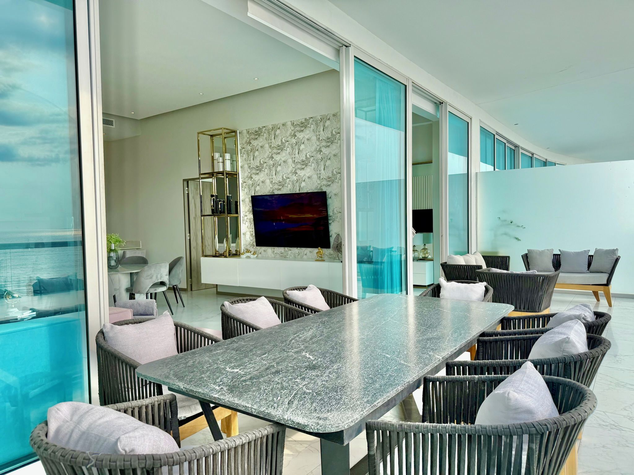 A modern interior with a dining area, wicker furniture, a TV mounted on the wall, and a view of the sea.