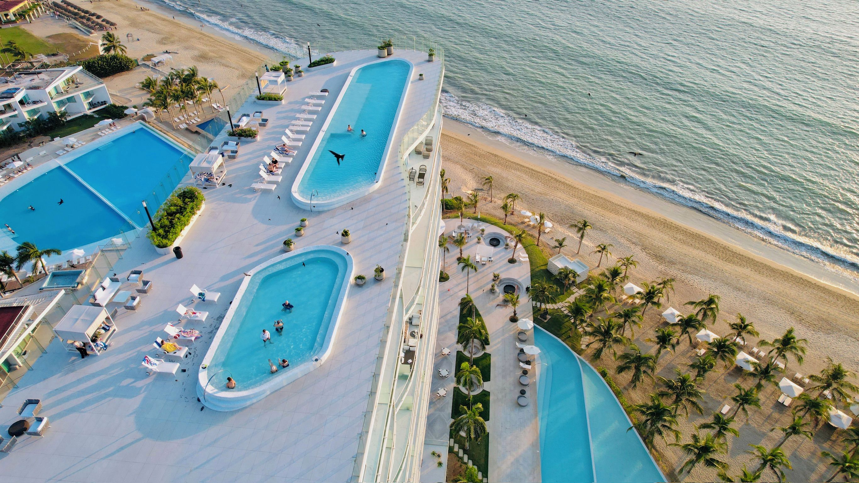 Eine Vogelperspektive zeigt einen luxuriösen Urlaubsort mit mehreren Swimmingpools, Zugang zum Strand und Palmen.