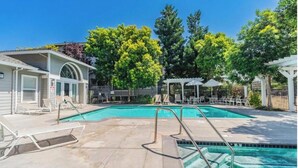 Una piscina con sillas de descanso, un área de estar techada y un edificio con acceso cubierto.