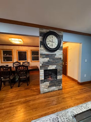 A living room with a stone fireplace, a dining area with wooden chairs, and a large wall clock.