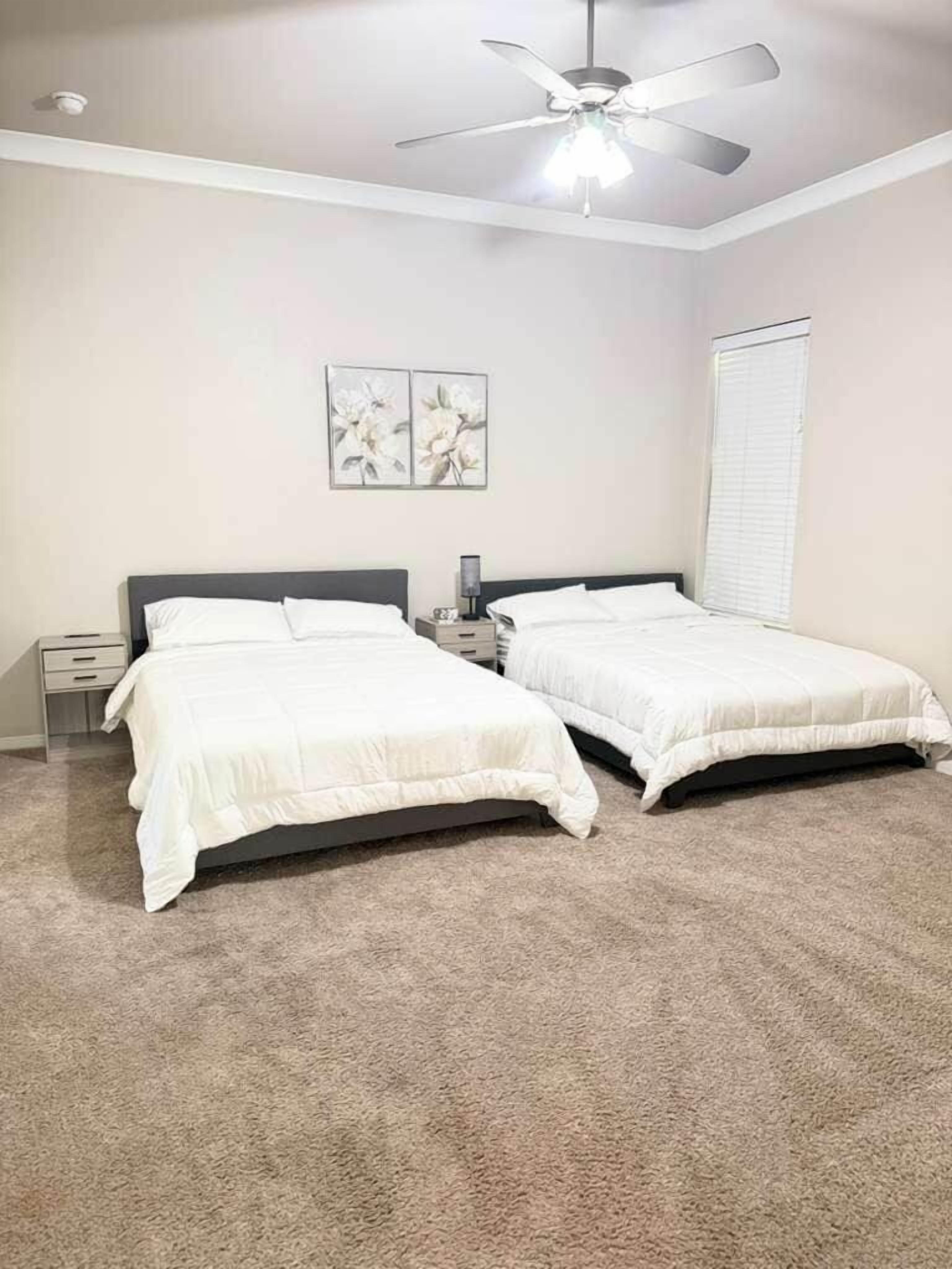 Une chambre avec deux lits simples, un ventilateur de plafond, une table de chevet avec une lampe et une fenêtre avec des stores.