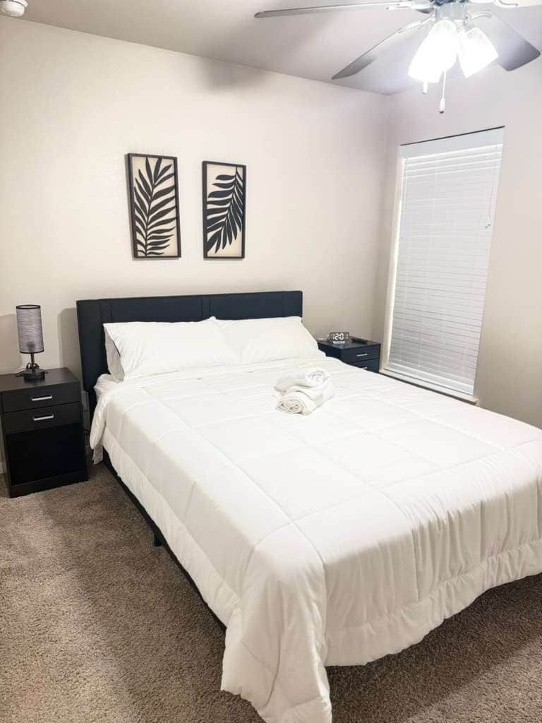 Een net opgemaakt bed in een kamer met een plafondventilator, een nachtkastje met een lamp en twee kunstwerken aan de muur.