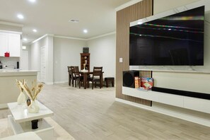 Un salon moderne comprenant un coin repas, un téléviseur fixé au mur et une petite étagère avec quelques objets.