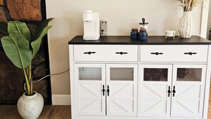 Un mobile bianco con maniglie nere, una macchina da caffè, un vaso con piante essiccate e due opere d'arte geometriche a parete.