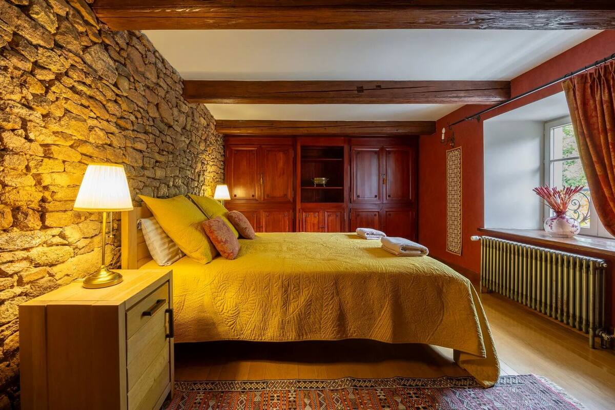 Un dormitorio con pared de piedra, vigas de madera, una cama con colcha amarilla, una mesita de noche con lámpara, una ventana con cortinas y un radiador.