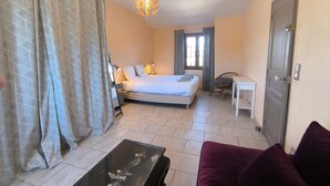 Une chambre d’hôtel comprenant un lit, un bureau, une chaise, un canapé, une table basse en verre, un rideau à motifs, un lustre et une porte.