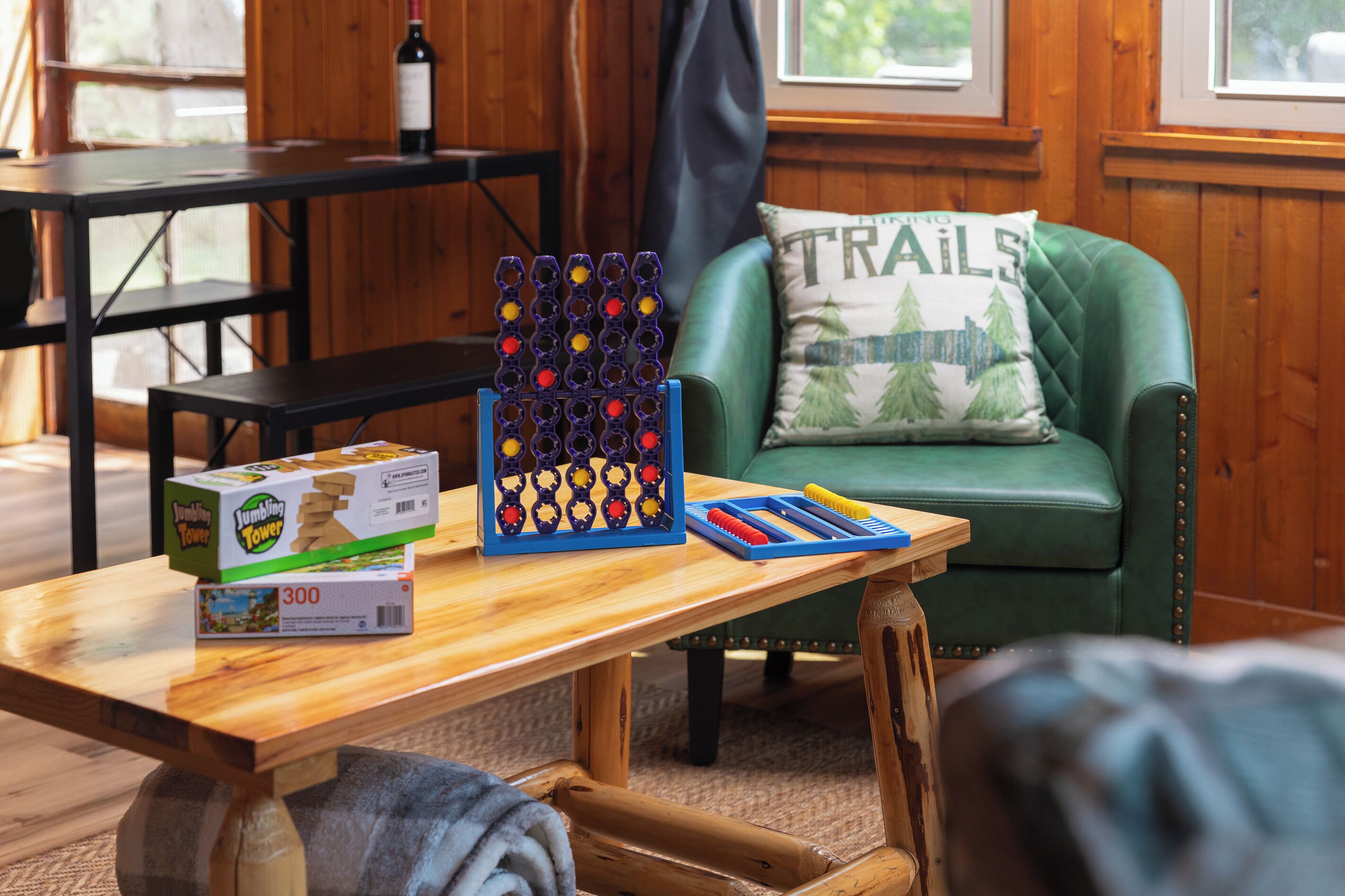 Una mesa de madera con un juego de torres JUMBLING TOWER, una silla verde con un cojín TRAILS y una botella de vino.