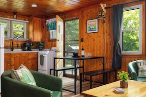 Interior acogedor de una cabaña con paredes de madera, una mesa de comedor, un sofá verde, una zona de cocina con estufa y cafetera, y vistas a los árboles a través de las ventanas.