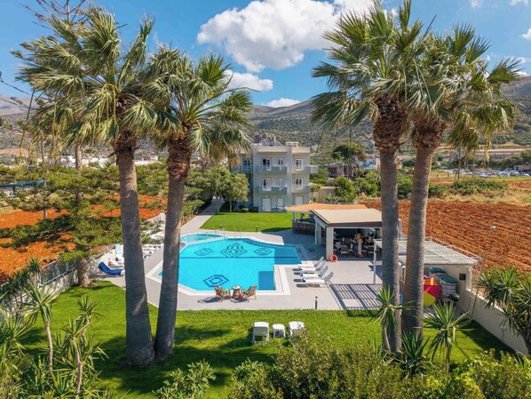 A pool with lounge chairs, palm trees, and a building with balconies. - Matthew's Place (Malia)