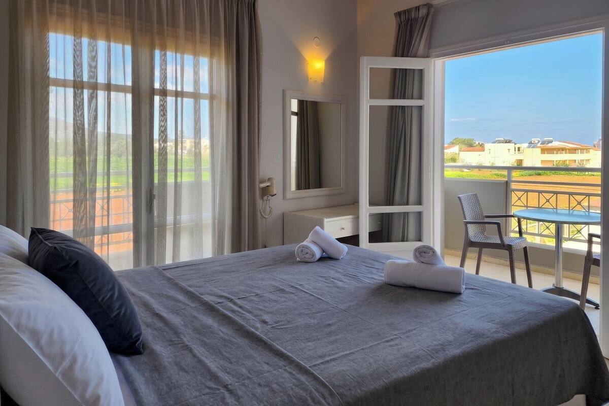 A hotel room with a bed, a desk, a chair, a balcony with a table, and a view of the landscape.