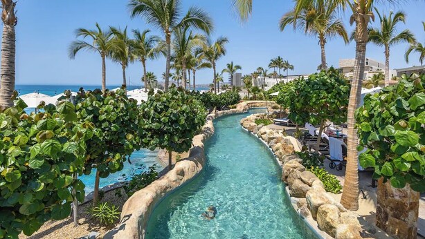 A clear water channel with palm trees and a pool on one side, surrounded by greenery and buildings. - Villa La Valencia (San José del Cabo)