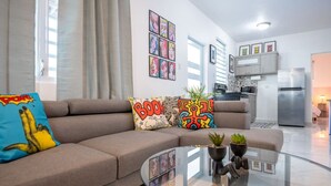 Een moderne woonkamer met een bank, kleurrijke kussens, een glazen salontafel en een keuken op de achtergrond.