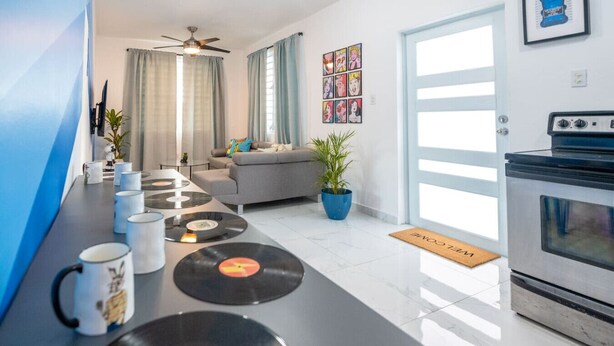 Een moderne keuken met een aanrecht waarop vinylplaten en mokken staan, een grijze bank, een eettafel en een groot raam met gordijnen.