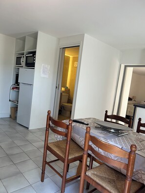 Ein Zimmer mit einem Bett, Holzstühlen, einer kleinen Küchenzeile inklusive Mikrowelle und Kühlschrank, sowie einem durchgehenden Türrahmen einsehbarer Badezimmer.