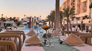 Un comedor con mesas dispuestas para dos, una piscina con toboganes y vistas a un edificio con balcones.