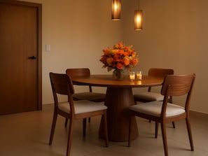 Un coin repas avec une table ronde en bois, quatre chaises, un vase de fleurs oranges, deux bougies et deux lampes suspendues.