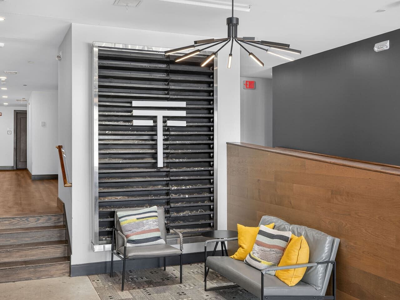 A modern lobby with a reception desk, a seating area with a sofa and chairs, a decorative wall with a large 'T' design, and a wooden accent wall.