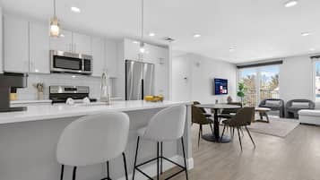A modern kitchen with white cabinetry, a central island with bar stools, a dining area with a table and chairs, and a large window providing natural light.