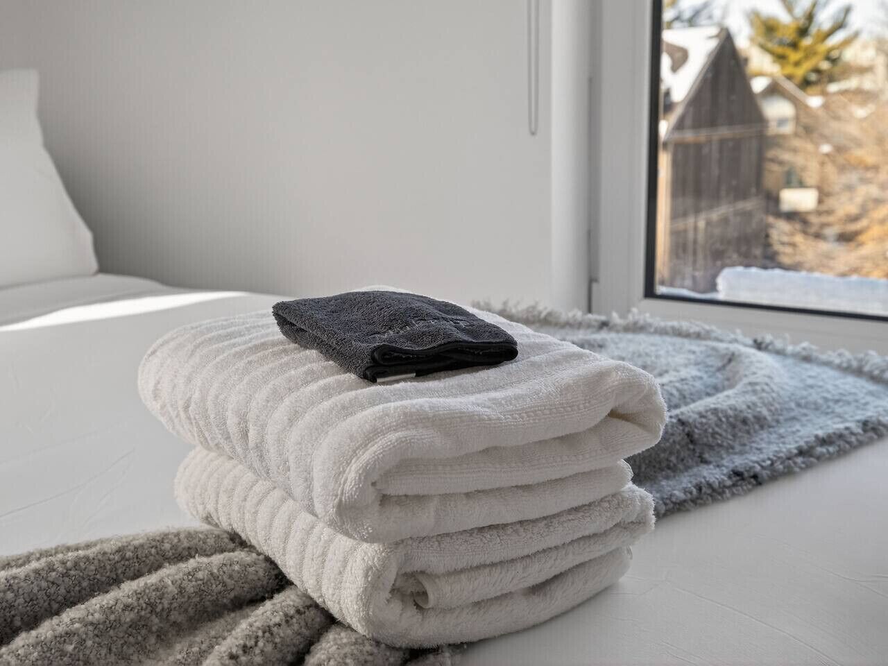 A neatly folded stack of white towels on a bed with a gray rug beside it, a window with a view of a building and trees, and a white wall.