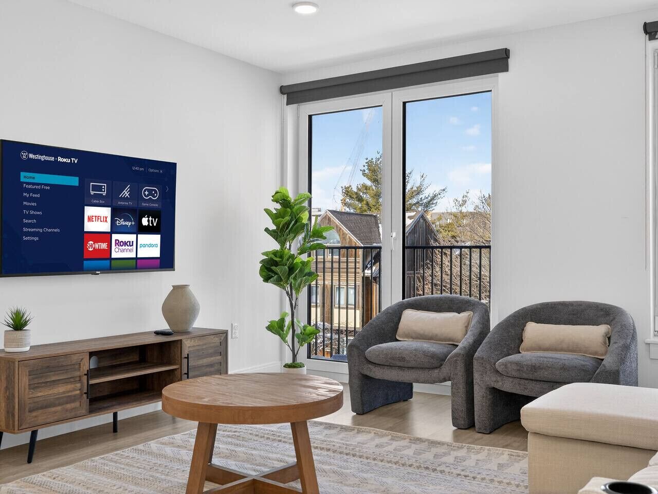 A modern living room with a flat-screen TV displaying a streaming service interface, a wooden coffee table, two grey armchairs, a small plant, and a view of a residential area through large windows.