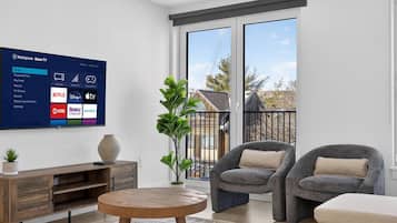 A modern living room with a flat-screen TV displaying a streaming service interface, a wooden coffee table, two grey armchairs, a small plant, and a view of a residential area through large windows.