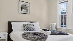 Cama bem arrumada com roupa de cama branca e cinza, um travesseiro preto e branco com bolinhas, uma obra de arte abstrata emoldurada acima da cama e uma janela com vista para prédios.