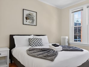 Una cama bien hecha con ropa de cama blanca y gris, un cojín blanco y negro con lunares, una obra de arte abstracta enmarcada sobre la cama y una ventana con vistas a edificios.