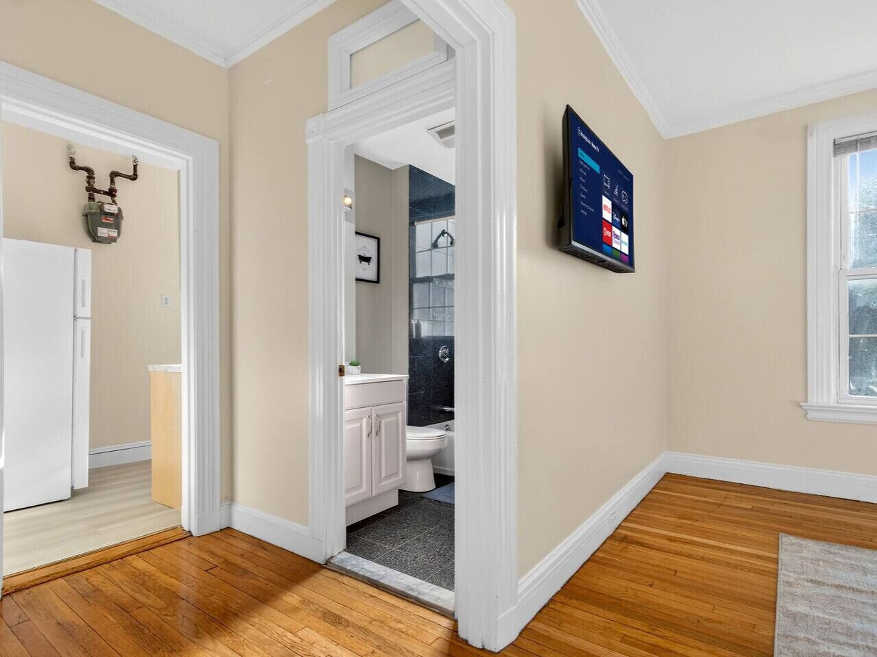 A hallway leading to a bathroom with a toilet, sink, and cabinet. There is a thermostat on the wall and a television mounted above the sink.