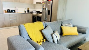 Una sala de estar moderna con un sofá gris, un cojín amarillo y almohadones estampados. Una mesa de centro de madera sostiene una caja. Se ve una cocina con armarios de madera y electrodomésticos de acero inoxidable al fondo.