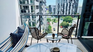 Un balcón con una mesa de vidrio, dos sillas de mimbre, una hamaca a rayas y vistas a edificios modernos.