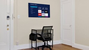 A room with a desk, a chair, a TV mounted on the wall displaying streaming services, and wooden flooring. - Allston 1BR Near Green Line | Sleeps 4 (Boston)