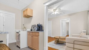A compact kitchen with a stove, oven, and wooden cabinets, leading to a living area with a sofa and a ceiling fan. - Allston 1BR Near Green Line | Sleeps 4 (Boston)