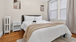 A bedroom with a bed, bedside table, radiator, and a window with curtains. - Allston 1BR Near Green Line | Sleeps 4 (Boston)