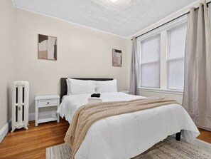 Une chambre à coucher avec un lit, une table de chevet, un radiateur et une fenêtre avec des rideaux.