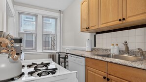 A kitchen with a stove, sink, wooden cabinets, and a window with blinds. - Allston 1BR Near Green Line | Sleeps 4 (Boston)